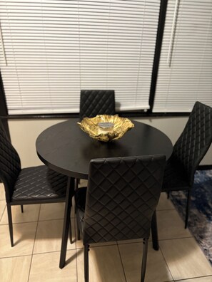 Ein runder Tisch mit schwarzem Tischtuch und einer goldenen Aschenbecher in der Mitte. Vier schwarze Stühle mit gesteppten Rückenlehnen stehen darum.
