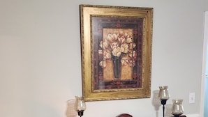 A living room with a wooden cabinet, a framed floral painting, and a small table with a candle. - Beautiful 3-bedroom house with AC in Elizabeth City (Elizabeth City)