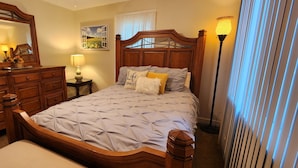 A bedroom with a wooden bed, a dresser, a nightstand with a lamp, a wall-mounted mirror, and a window with curtains. - Beautiful 3-bedroom house with AC in Elizabeth City (Elizabeth City)
