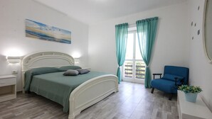 A bedroom with a bed, a chair, a nightstand, a window with curtains, and a painting on the wall. - Lovely apartment in Castellabate (Castellabate)