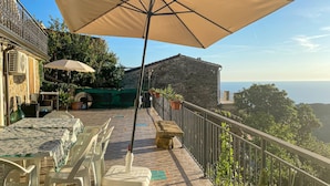 Terraço ao ar livre com mesa posta para refeições, um grande guarda-sol e vista para o mar.