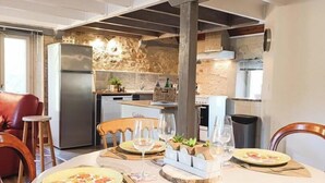 Un coin repas avec une table ronde dressée pour deux personnes, une cuisine en arrière-plan et un mur en pierre.