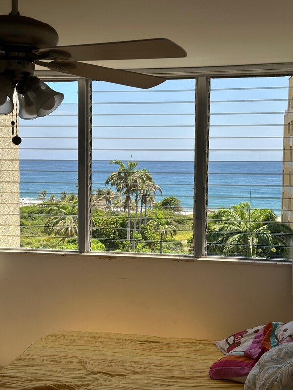 Ein Schlafzimmer mit Bett, Deckenventilator und großem Fenster mit Blick auf das Meer und Palmen.