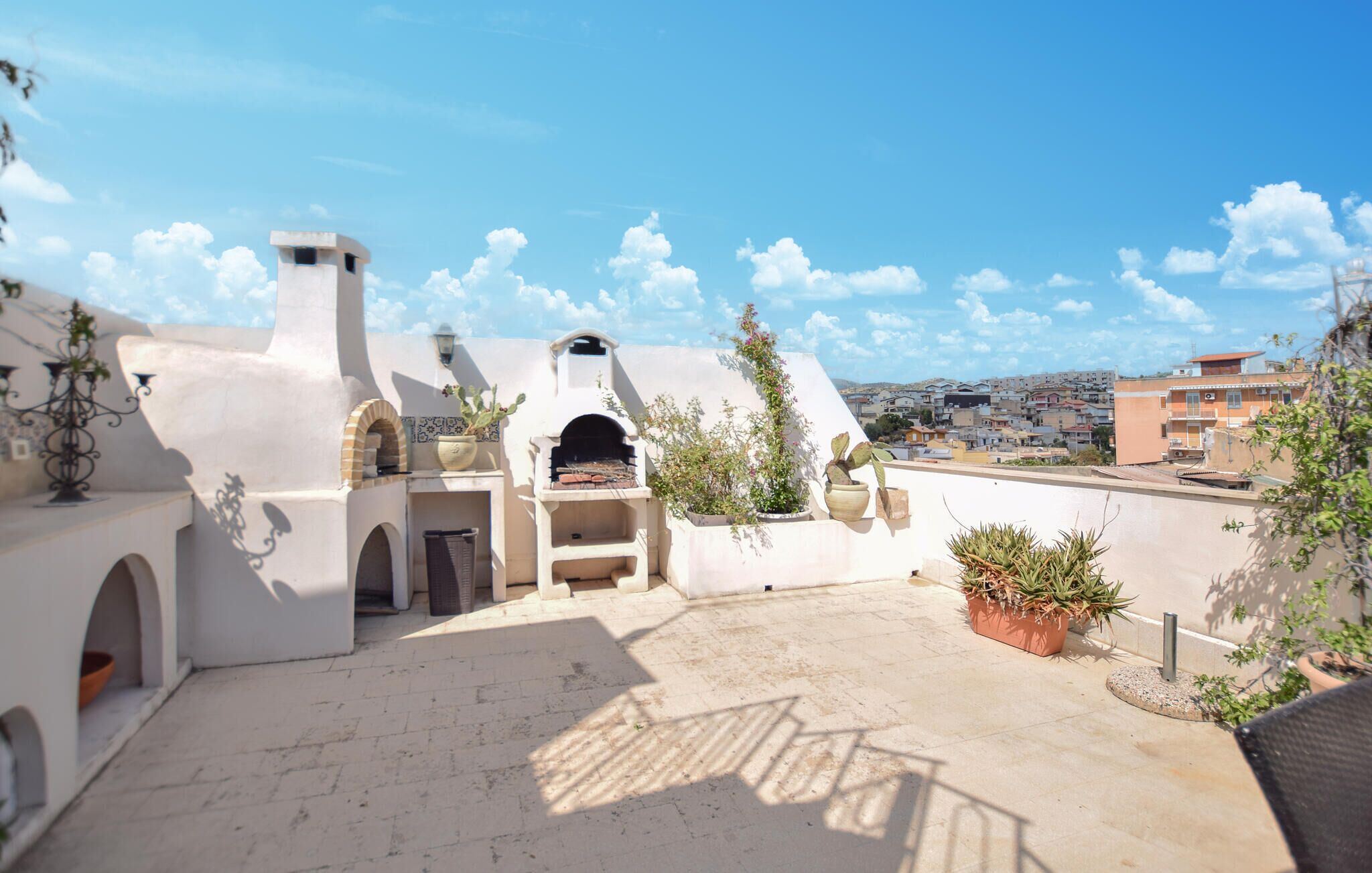 Terraza en la azotea con área de barbacoa, plantas en macetas y vistas de la ciudad.