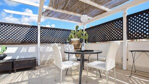 An outdoor patio with a round table and white chairs, a cactus plant, and a pergola with a woven roof.