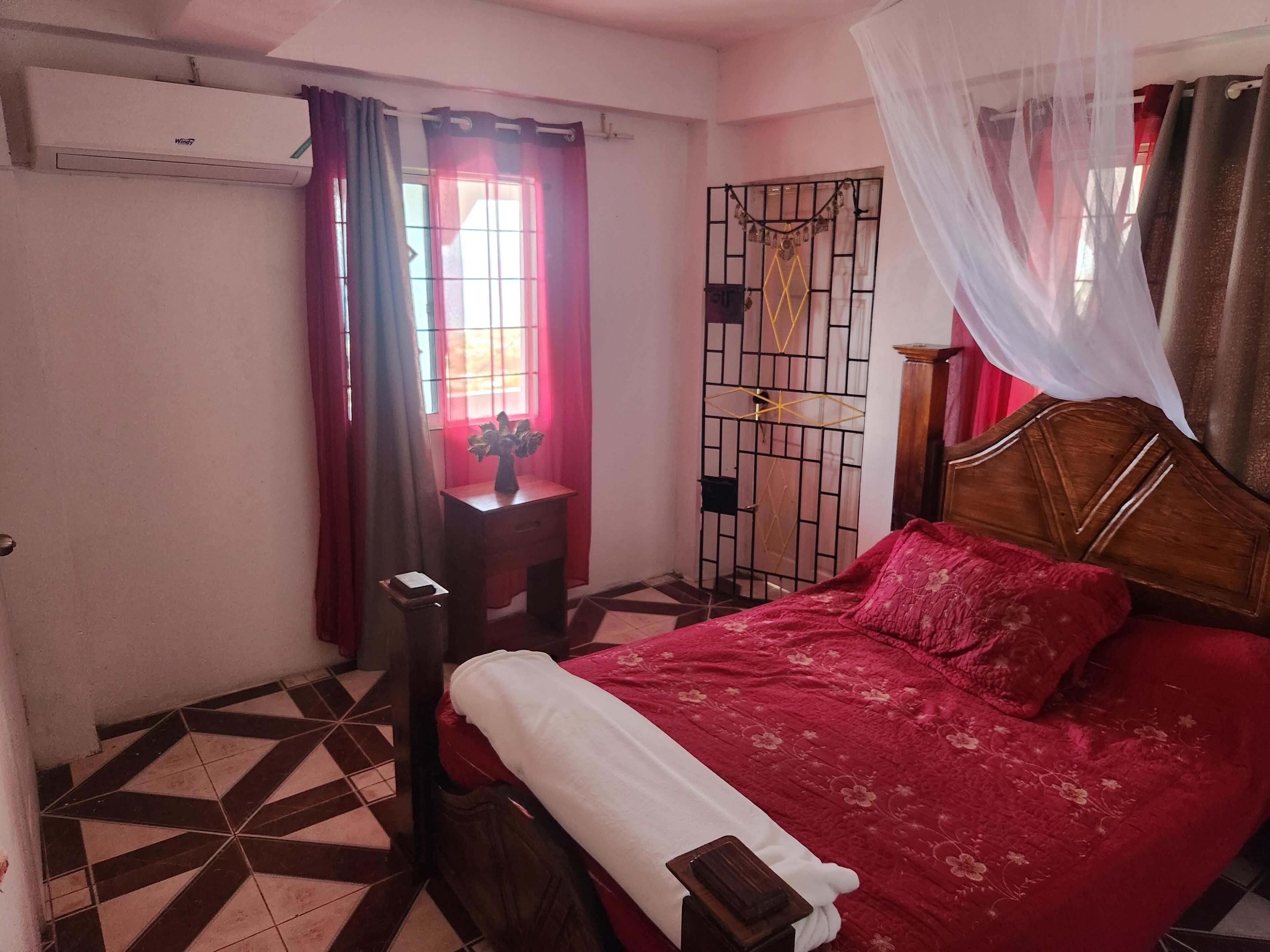 Une chambre à coucher comprenant un lit, une table de chevet, une fenêtre avec des rideaux, une porte avec un portail en métal et un appareil de climatisation.