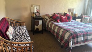 A bedroom with a wooden chair, a bed with plaid bedding, a nightstand with a clock, and a framed picture on the wall.