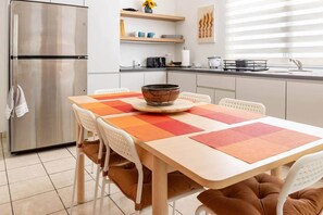 Une cuisine moderne avec un réfrigérateur en acier inoxydable, une table à manger agrémentée d’un set de table coloré, des chaises blanches et un banc marron avec un coussin.