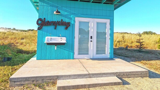 A small, turquoise building with a sign reading "Glamping." It features a white door, a small sink, and a metal sign. - Vinyl Vineyards  (Paso Robles)