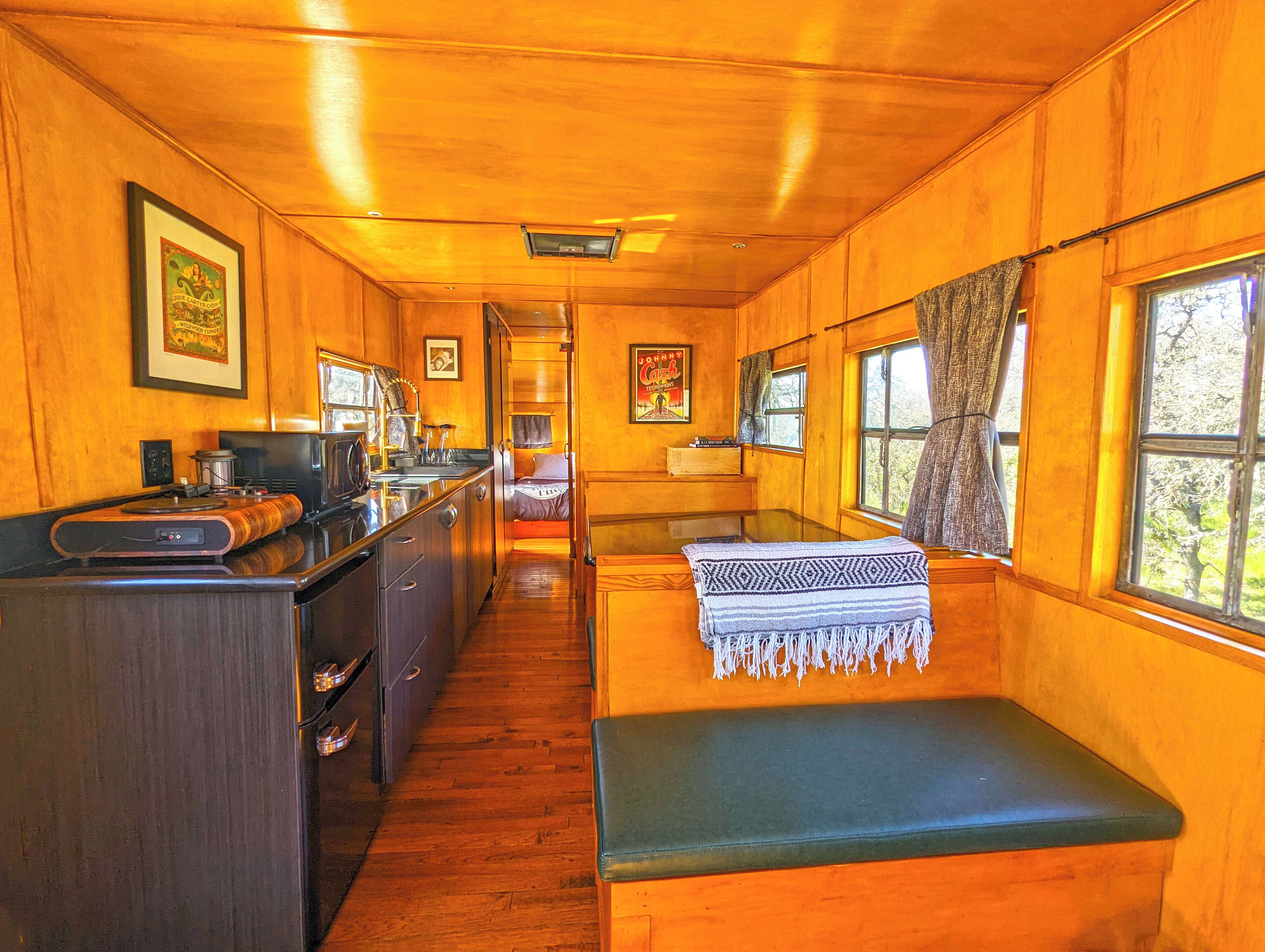 A cozy cabin interior with wooden walls and floors, a kitchenette, a dining area, and a bench with a blanket.