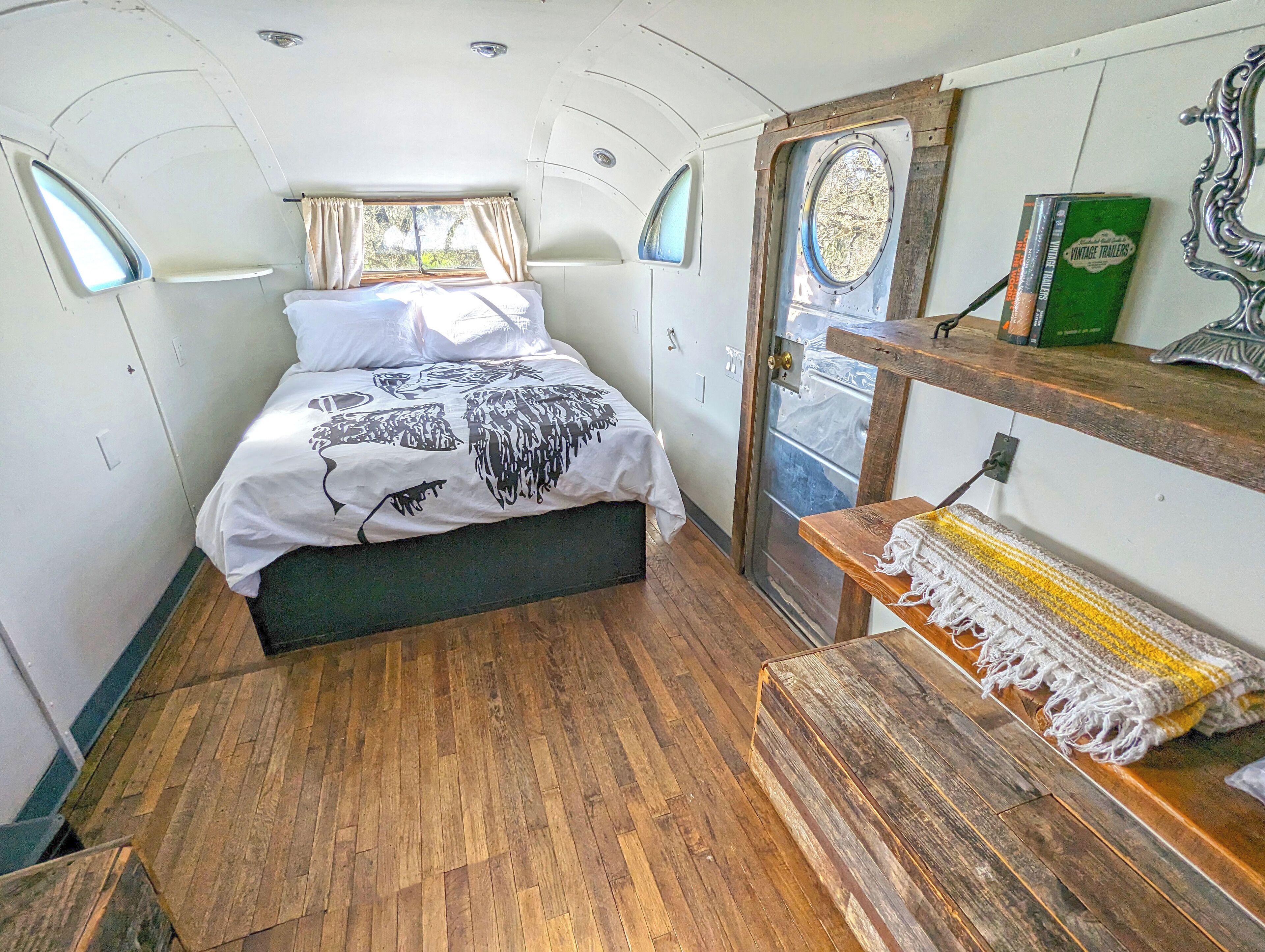 Une pièce compacte et bien éclairée, avec un lit, un plancher en bois, un petit banc et une porte donnant sur l’extérieur.