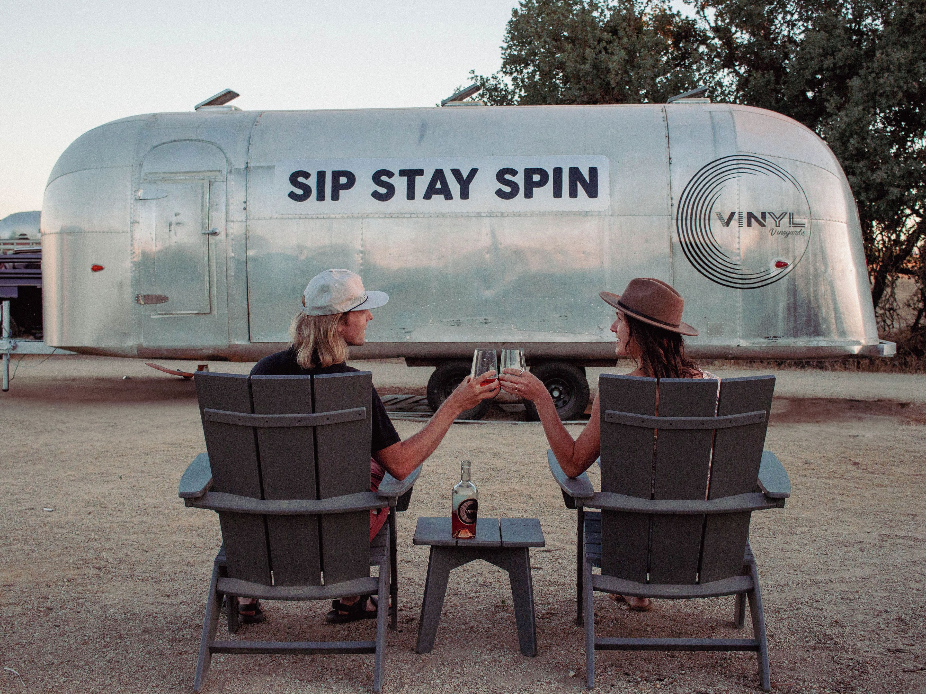 Twee personen zitten aan een klein tafeltje met stoelen, gericht op een vintage caravan met de tekst 'SIP STAY SPIN' en een vinyl logo.