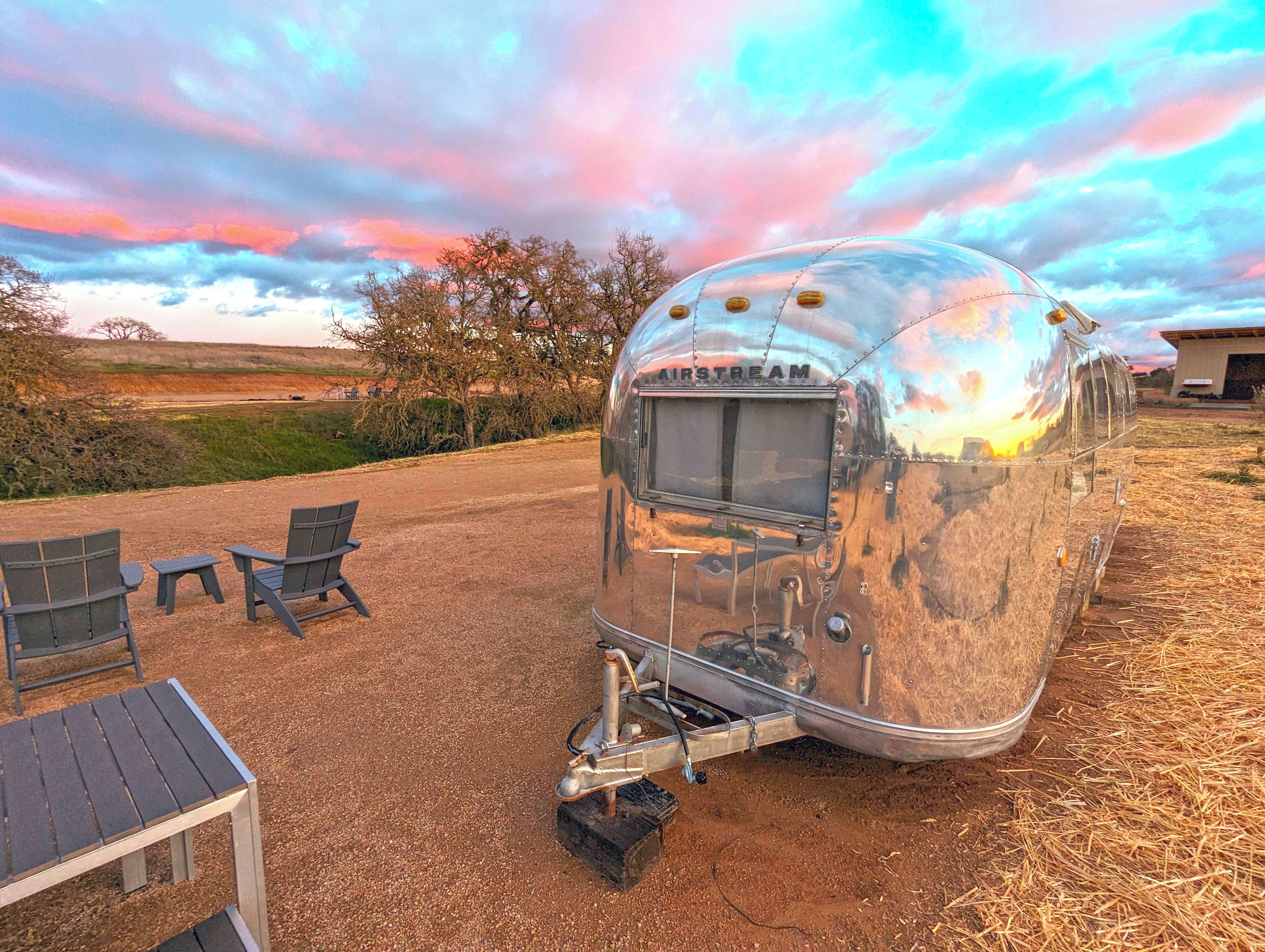 A vintage trailer with a flatbed, two chairs, and a small table in a rural setting.
