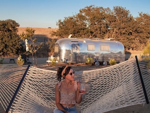 A person sitting on a hammock with a vintage trailer in the background.
