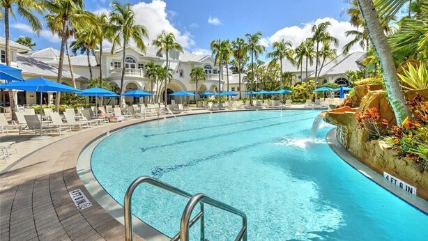 A large swimming pool with lounge chairs and umbrellas, surrounded by palm trees and a building with a white facade. - Beautiful First-floor, End Unit, Coach Home in Fiddler's Creek, Naples, FL (Naples)