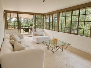 Ein großzügiger Raum mit einem großen Fenster, einem Sofa, einem Glastisch und einem Essbereich, der durch das Fenster sichtbar ist.