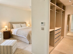 Une chambre à coucher comprenant un lit, des tables de chevet, une commode, un pouf rayé et un dressing avec des tiroirs, ainsi qu’un thermostat fixé au mur.