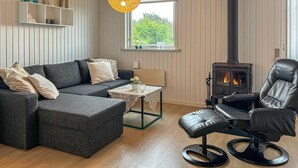 A cozy living room with a grey sectional sofa, a black leather chair, a small table with a lace tablecloth, and a wood-burning stove.