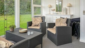 A room with wicker furniture, a small table with a bowl and glasses, and a view of a green garden through large windows.