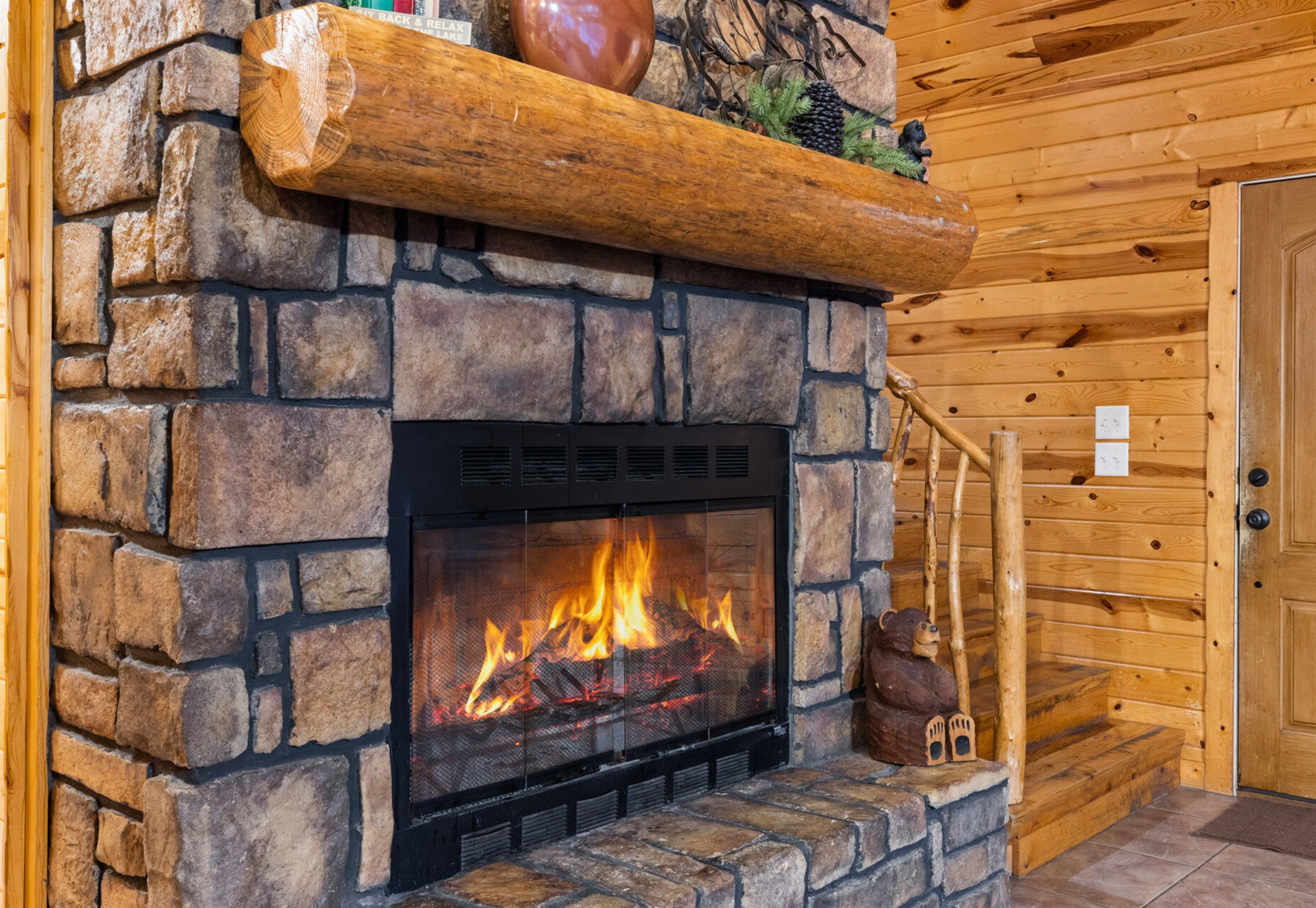 Une cheminée en pierre avec un feu de bois, des bûches et une étagère rustique agrémentée d’objets décoratifs.