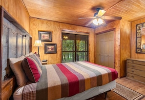 Un dormitorio con paneles de madera, una cama, una mesita de noche, un aparador y una ventana con persianas.