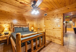 A wooden cabin room with a bed, a ceiling fan, and a nightstand with a lamp. - Trails | Fire Pit | 4 Stocked Ponds | Hot Tub (Broken Bow)