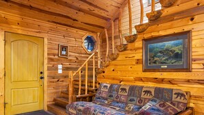 A wooden cabin interior with a yellow door, a staircase, a sofa with animal-themed cushions, a framed picture of a fish, and a small window. - Shaui Chukka Cabin | Cozy Pondside Cabin (Broken Bow)