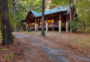 A rustic lodge with a covered porch and stone pillars, surrounded by a wooded area. - White Tail Waters | Romantic Pond-View Cabin (Broken Bow)