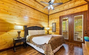 A wooden-paneled bedroom with a bed, nightstands, a ceiling fan, and a door leading to an outdoor area. - White Tail Waters | Romantic Pond-View Cabin (Broken Bow)