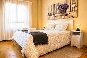 A bedroom with a bed, nightstands, a bedside lamp, a rug, a window with curtains, and a wall art piece.