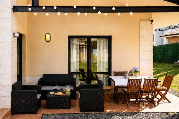 An outdoor patio with a sofa, dining table, and chairs, a small table with a fruit bowl, and a view through a glass door.
