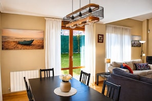 A dining area with a table, chairs, a vase of flowers, a painting of a boat, and a view of a garden through a glass door.