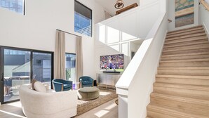 A spacious living area with a curved white sofa, a round ottoman, and a blue chair. A wooden staircase leads to an upper level with a large window and a painting.