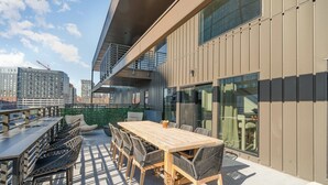 An outdoor terrace with a wooden table and chairs, a glass wall, and a view of the cityscape.