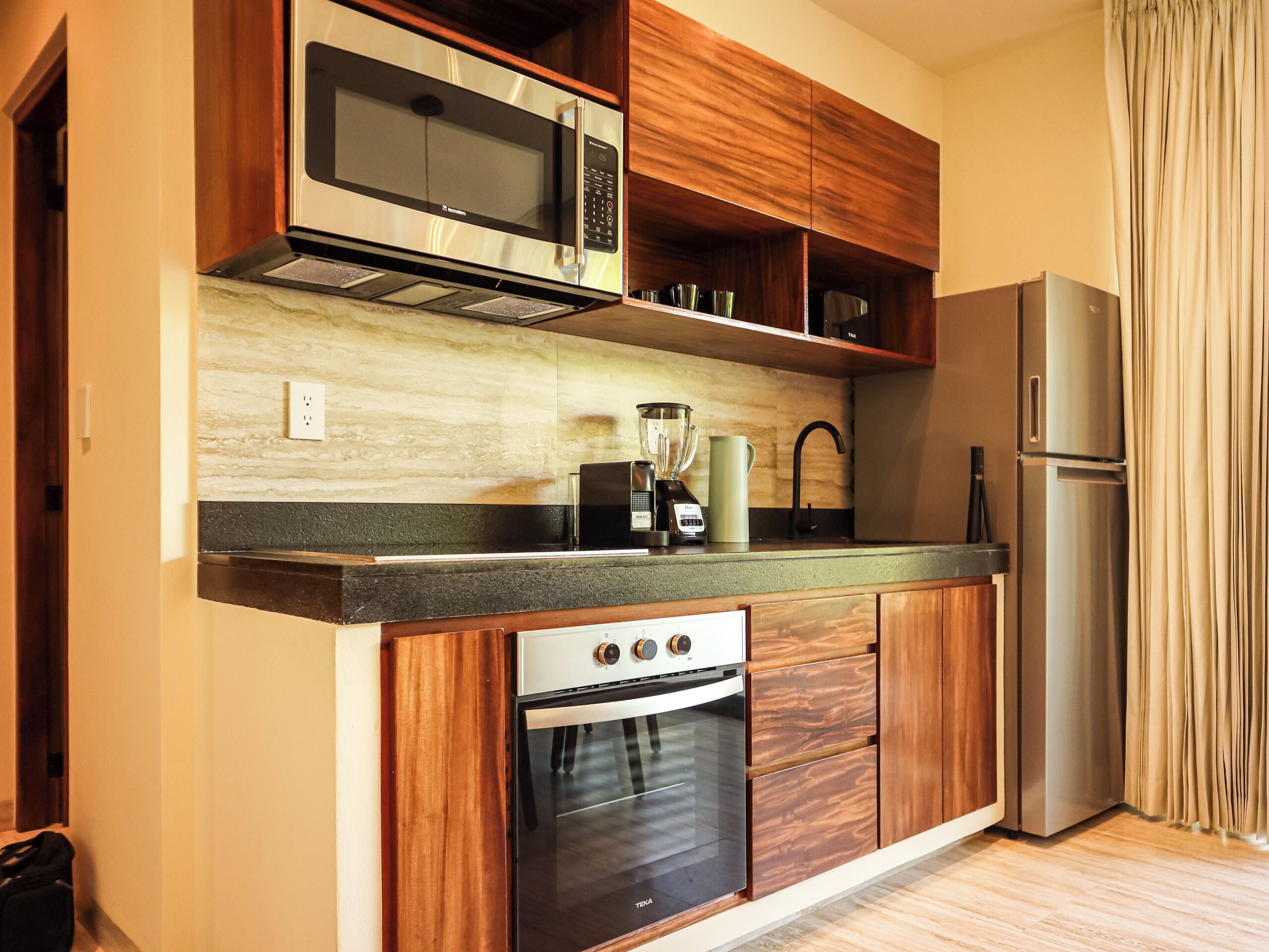A compact kitchen with a built-in microwave, a blender, a kettle, and a refrigerator.