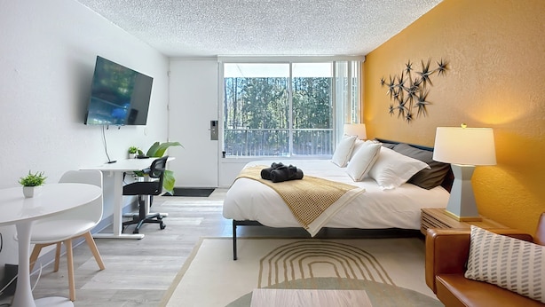 A hotel room with a bed, a desk, a chair, a TV, and a balcony with a view of trees. - Cozy King Studio | Zoo, DT & Jacksonville U (Jacksonville)