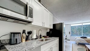 A modern kitchen with white cabinets, a granite countertop, a microwave, a coffee maker, and a refrigerator. - Cozy King Studio | Zoo, DT & Jacksonville U (Jacksonville)