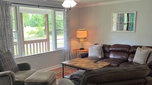 A living room with a brown leather sofa, a matching armchair, and a small table with a lamp. A large window with a view of greenery is visible.