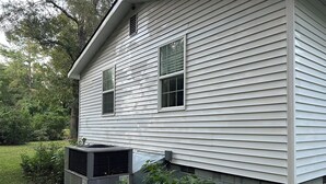 A house with white siding, two windows, and an air conditioning unit on the ground.