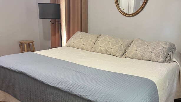A neatly made bed with a grey and white quilt, a wooden floor, a small stool, and a lamp.