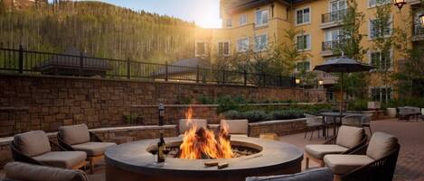 A circular fire pit surrounded by wicker chairs and tables, set against a backdrop of a multi-story building with balconies and a stone wall.