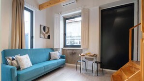 Une pièce avec un canapé bleu, une petite table avec des tabourets, une fenêtre avec des rideaux et une porte noire.