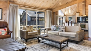 Una sala de estar acogedora con sofá, sillón, mesa de centro, techo de madera, ventanas amplias y una zona de cocina.