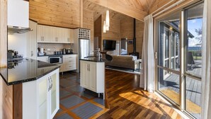 Una cabaña de troncos moderna con una cocina que cuenta con gabinetes blancos, una encimera negra y una isla central. Hay un amplio ventanal corredero que da a una terraza exterior.