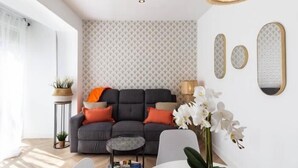 Een moderne woonkamer met een grijze bank, oranje kussens, een klein rond tafeltje en een plant in pot.