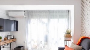 Een moderne woonkamer met een grijze bank, een klein bijzettafeltje met een plant, een wit gordijn en een wandgemonteerde televisie.