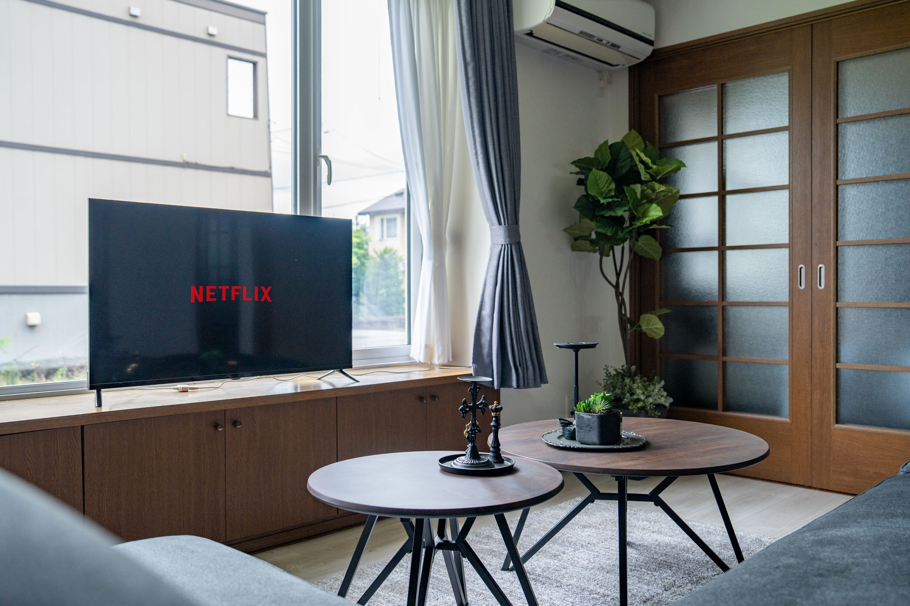 Uma sala de estar moderna com uma televisão de tela plana em um armário de madeira, uma mesa redonda com um abajur preto e uma planta em vaso.