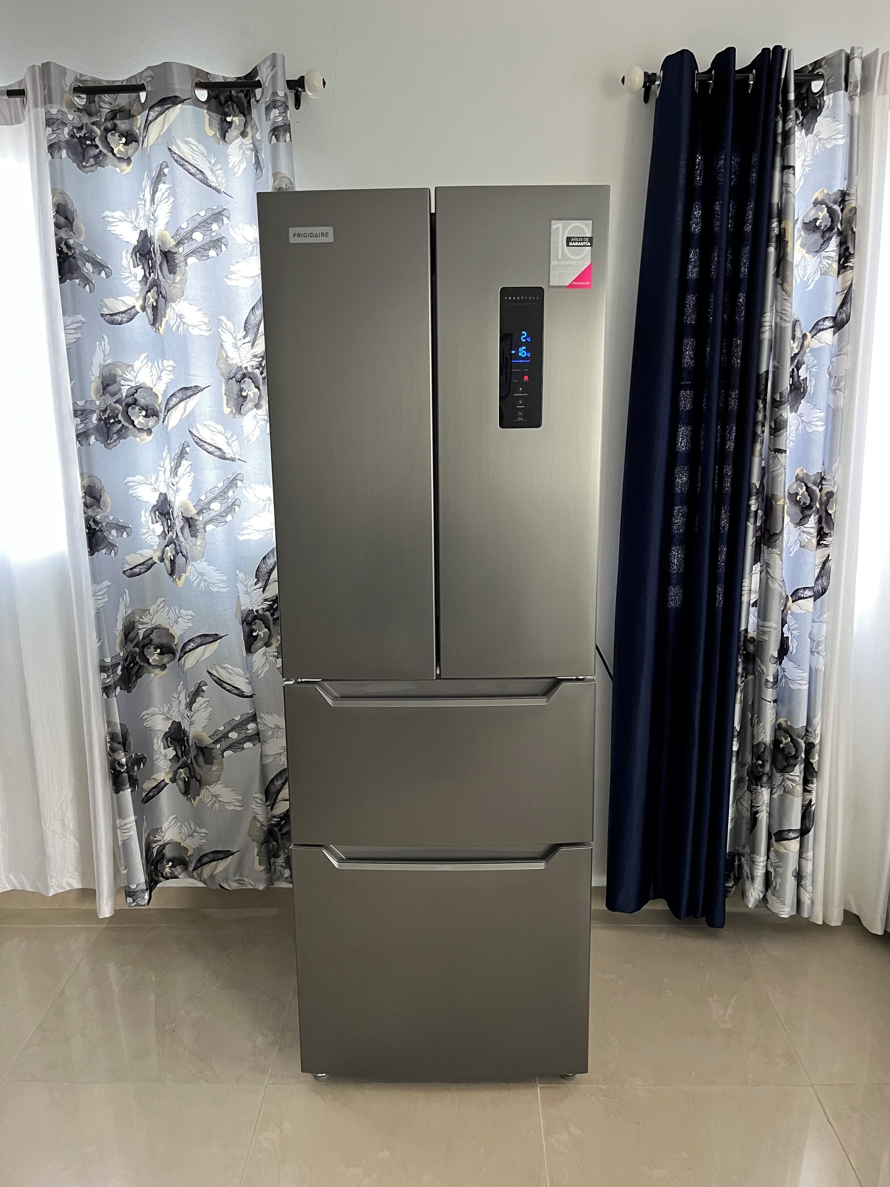 Ein Edelstahl-Kühlschrank mit digitaler Anzeige, in einem Raum mit blumigem Vorhang und Fliesenboden.