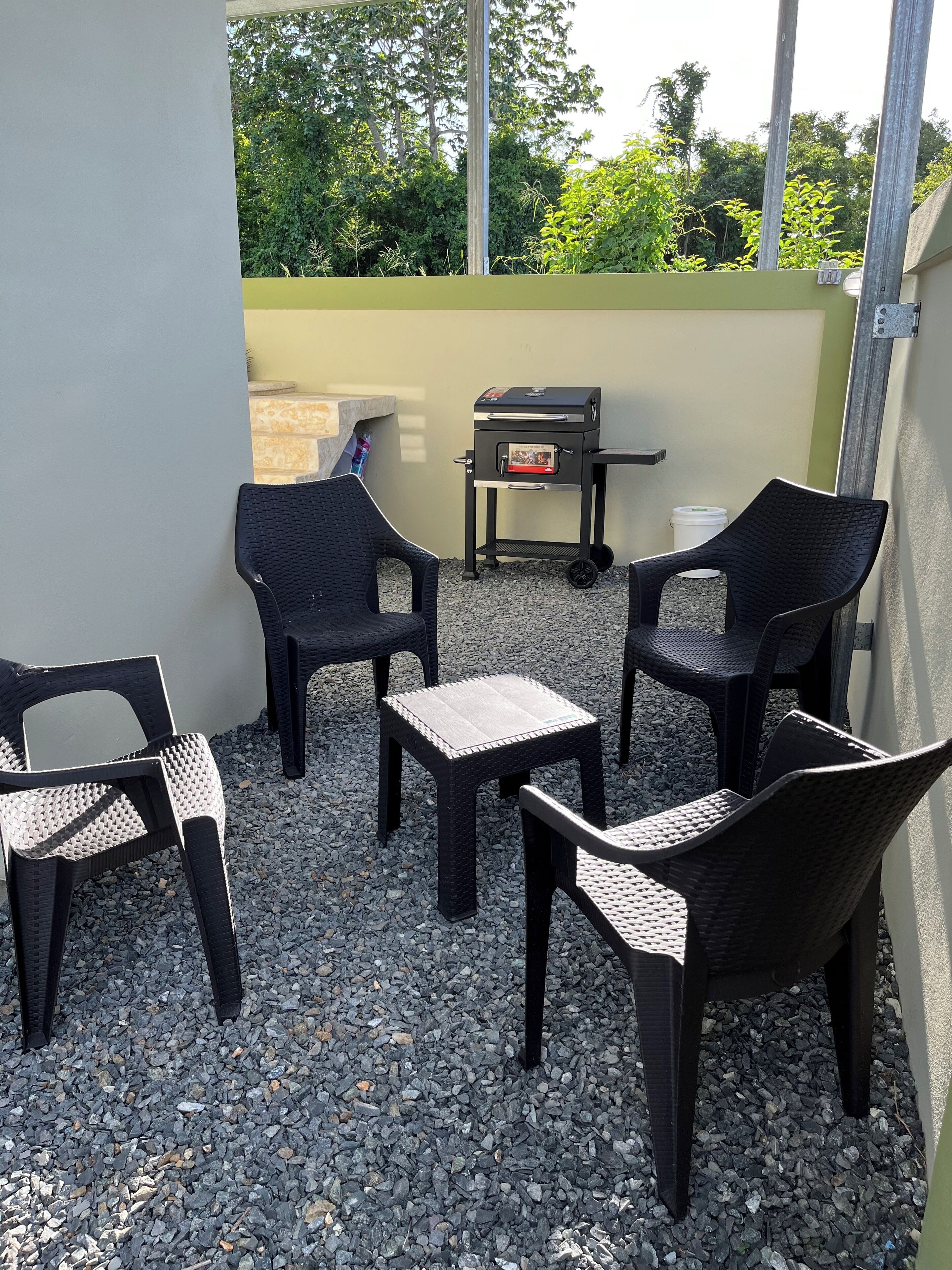A patio area with wicker chairs and a small table, a barbecue grill, and a view of greenery.