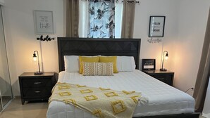Ein ordentlich bezogenes Bett mit weißer Daunendecke und einer dekorativen Gold-Weiß-Decke. Zwei Nachttische mit Lampen, ein gerahmter Motivationsspruch und ein gerahmtes Bild an der Wand.